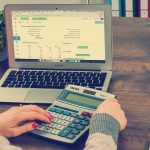 Mains sur une calculatrice devant un ordinateur portable, calcul d'un budget netlinking