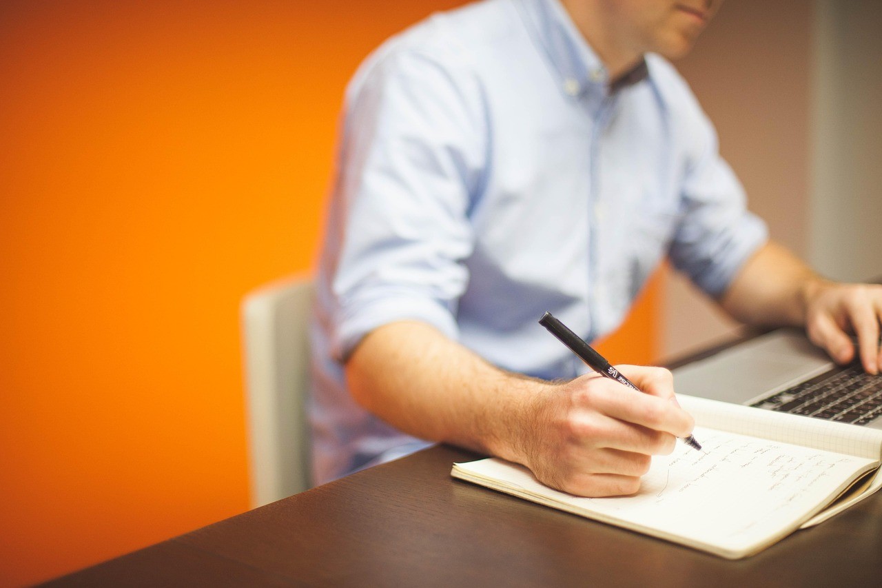 Personne écrivant à la main dans un carnet à côté d'un ordinateur portable, sur un bureau